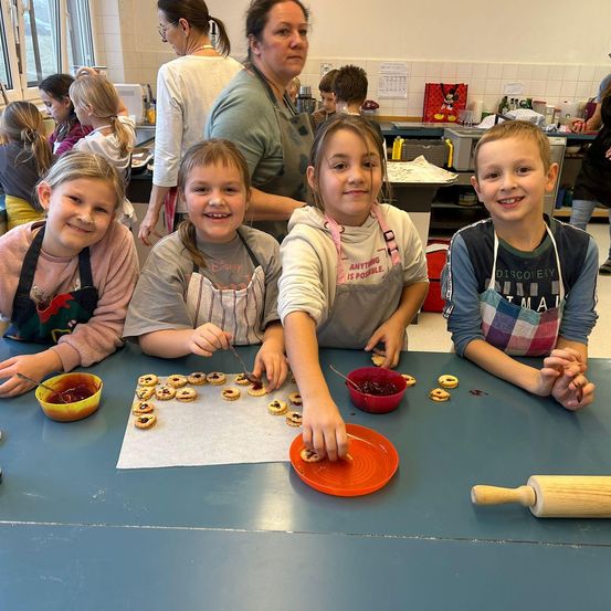 Mehrere Kinder sitzen in einem Klassenzimmer um einen Tisch herum und beschäftigen sich mit einer Backaktivität. Sie tragen Schürzen und haben verschiedene Backutensilien und Zutaten vor sich. Die Kinder lächeln und scheinen die Aktivität zu genießen. Im Hintergrund beaufsichtigen einige Erwachsene.