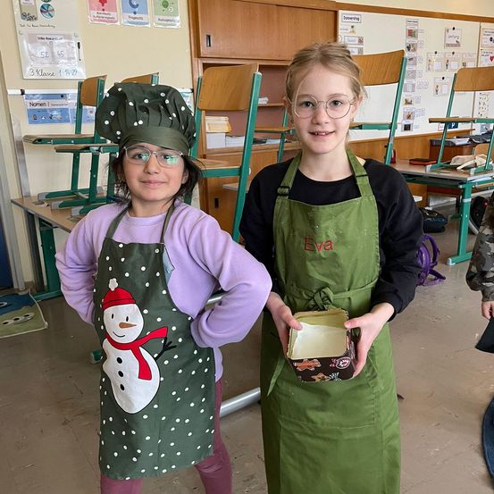 Zwei junge Mädchen in einem Klassenzimmer, beide in Schürzen. Eine trägt eine Kochmütze und einen Schneemann auf ihrer Schürze, die andere hält eine Box. Hinter ihnen sind gestapelte Stühle und eine Tafel.