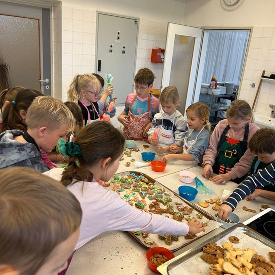 Eine Gruppe von Kindern verziert Lebkuchen in einem Klassenzimmer. Sie sind von Schüsseln und Tabletts mit Keksdekorationen umgeben.