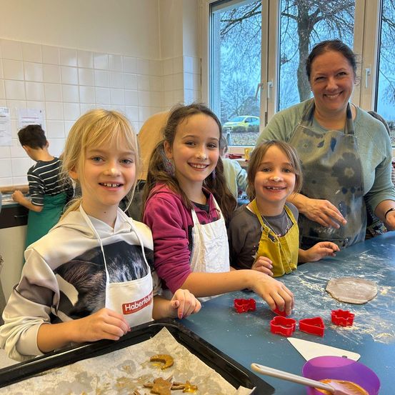 Drei Kinder in Schürzen lächeln und verwenden Ausstechformen auf einem blauen Tisch, eine Frau im Hintergrund.
