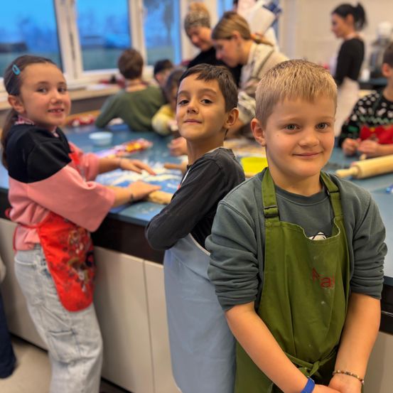 Drei Kinder in Schürzen lächeln in die Kamera. Sie stehen vor einem Tisch mit verschiedenen Gegenständen darauf. Hinter ihnen arbeiten andere Kinder und Erwachsene an einem Projekt. Der Raum hat große Fenster und es liegt ein Nudelholz auf dem Tisch.