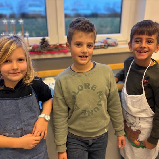 Drei Kinder, zwei Jungen und ein Mädchen, stehen zusammen in einem Raum mit einem festlich dekorierten Fenster im Hintergrund. Sie tragen Schürzen und schauen zur Kamera.