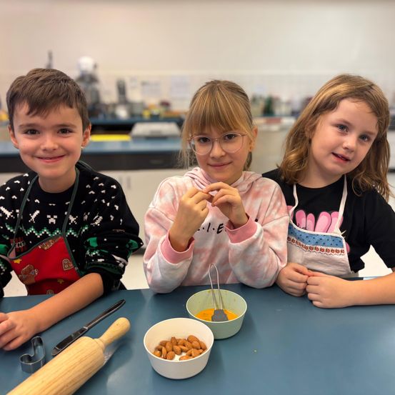 Drei Kinder, zwei Mädchen und ein Junge, sitzen an einem Tisch, wahrscheinlich in einem Klassenzimmer. Sie tragen Schürzen und scheinen ein Rezept zuzubereiten. Auf dem Tisch stehen eine Schüssel mit Mandeln, ein Nudelholz und ein Messer. Dahinter ist ein unscharfer Hintergrund eines Tresens mit verschiedenen Gegenständen.