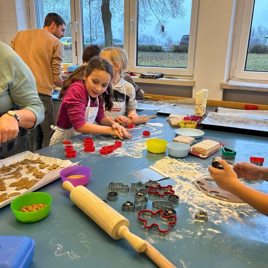 Eine Gruppe von Kindern und Erwachsenen backt Kekse in einem Klassenzimmer. Sie verwenden Ausstechformen und Nudelrollen. Der Tisch ist mit Keksteig und verschiedenen Ausstechformen gefüllt.