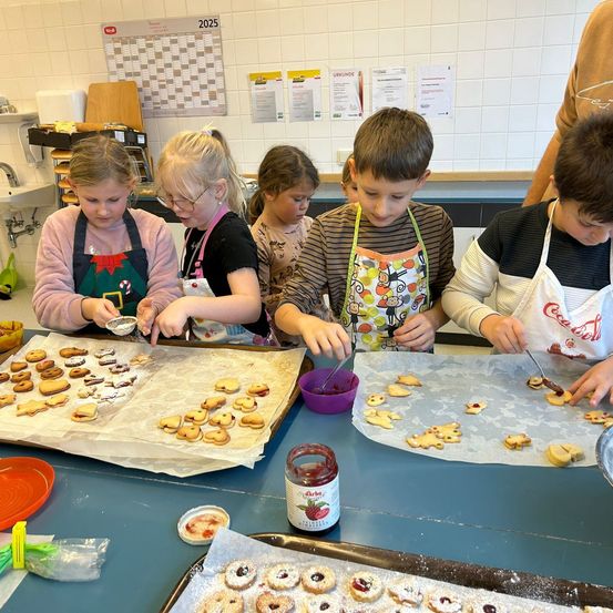 Mehrere Kinder verzieren Kekse mit Fruchtaufstrich in einer Küche. Sie tragen Schürzen und konzentrieren sich auf ihre Aufgabe. Es gibt Tabletts mit Keksen, Schüsseln mit Marmelade und andere Küchenutensilien auf dem Tisch.
