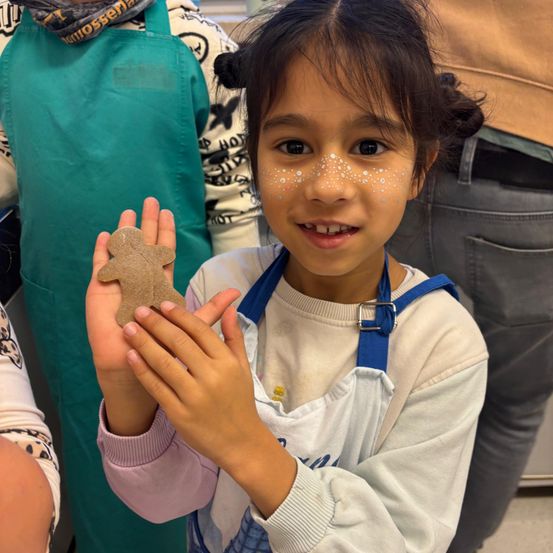 Ein junges Mädchen mit Gesichtsbemalung hält einen Lebkuchenmann und lächelt. Sie trägt einen weißen Pullover mit einer blauen Schürze. Hinter ihr steht eine Person in einer grünen Schürze, und eine weitere in grauer Jeans ist teilweise sichtbar.