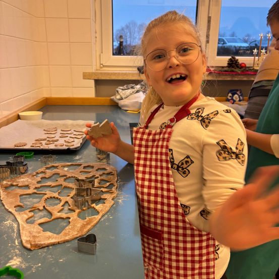 Ein junges Mädchen mit Brille und einer Schürze steht mit einem Lächeln im Gesicht in einer Küche. Sie hält einen Keks in der Hand. Die Küche hat eine Arbeitsplatte, ein Tablett und Ausstechformen. Im Hintergrund befindet sich ein Fenster mit Blick auf ein Auto und Bäume.