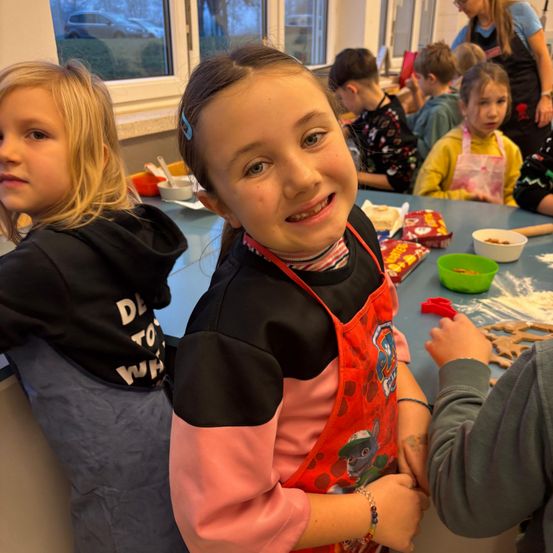 Mehrere Kinder in einem Klassenzimmer bereiten Kekse zu. Ein Mädchen in einer roten Schürze lächelt in die Kamera, während andere im Hintergrund stehen.
