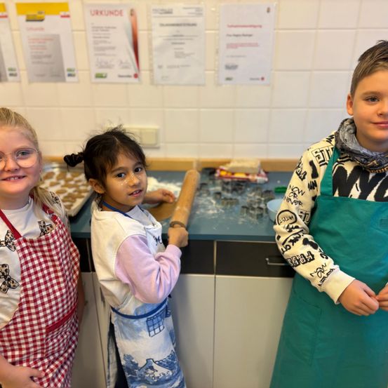 Drei Kinder in Schürzen stehen lächelnd in einer Küche. Ein Mädchen benutzt einen Nudelholz auf einem Tisch mit Keksen und Ausstechern.