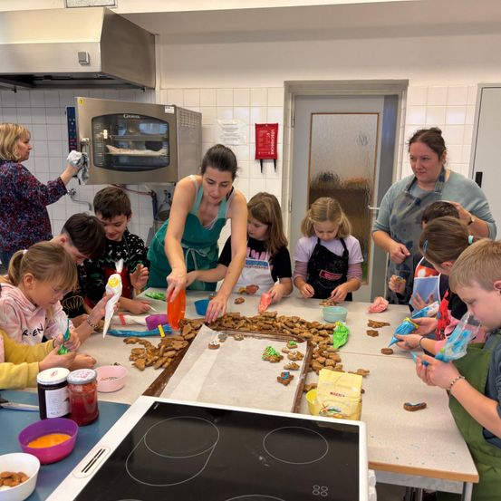 Eine Gruppe von Kindern dekoriert Kekse in einer Küche unter Aufsicht von Erwachsenen. Schüsseln, Gläser und Keksbleche stehen auf dem Tisch.