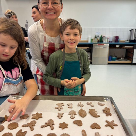 Eine Frau und zwei Kinder sind in einer Küche, tragen Schürzen und lächeln. Sie stehen vor einem Tablett mit gebackenen Lebkuchen. Die Frau hält ein Tablett mit Keksen darauf.
