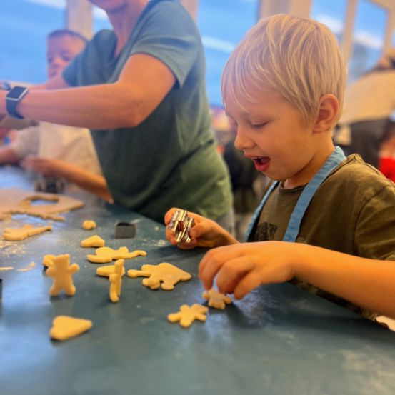 Ein junger Junge in einem braunen Shirt macht mit Ausstechern Lebkuchenmänner am Tisch. Hinter ihm macht eine Frau auch Lebkuchenmänner.