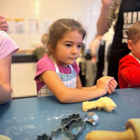 Ein kleines Mädchen sitzt an einem Tisch, trägt ein rosa T-Shirt und eine Schürze, umgeben von Ausstechformen und Teig. Sie konzentriert sich auf ihre Aufgabe. Dahinter sind verschwommen eine Person in einem roten Shirt und eine weitere in einer schwarzen Schürze zu sehen.