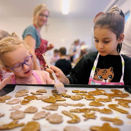 Zwei junge Mädchen dekorieren Lebkuchen in einer Küche. Sie verwenden einen Pinsel, um eine goldene Flüssigkeit aufzutragen. Andere Menschen sind im Hintergrund.