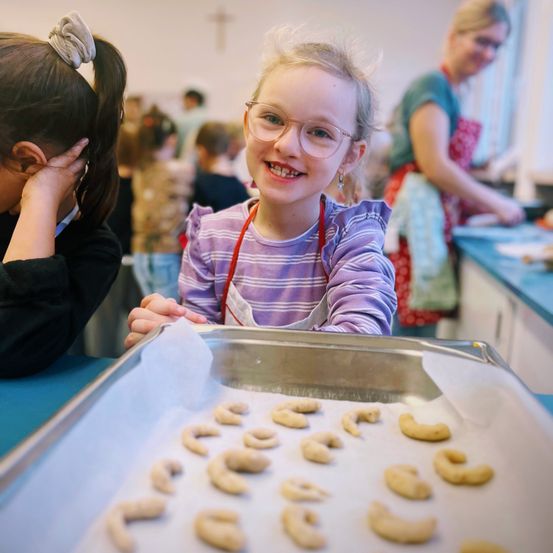 Ein junges Mädchen mit Brille lächelt in die Kamera, umgeben von Keksen auf einem Tablett. Ein weiteres Kind und eine Frau im Hintergrund sind ebenfalls an der Backaktivität beteiligt.