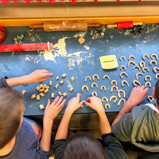 Vier Kinder sitzen am blauen Tisch und ordnen Keksteigstücke an, um Kekse zu backen. Kekse und Ausstechformen sind auf dem Tisch verstreut.