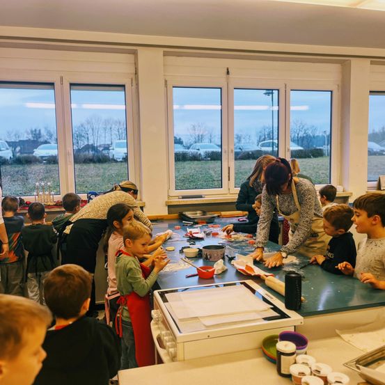 Kinder versammeln sich um einen Tisch in einer Küche, wobei Erwachsene sie anleiten. Sie tragen Schürzen und scheinen an einer Aktivität beteiligt zu sein. Der Raum hat große Fenster mit Blick auf ein Grasfeld.
