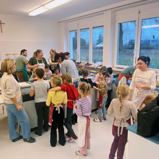 Eine Gruppe von Erwachsenen und Kindern in einer Küche, einige mit Schürzen. Sie scheinen an einer Kochaktivität teilzunehmen, wobei Erwachsene hinter einer Theke stehen und Kinder um sie herum versammelt sind. Der Raum ist gut beleuchtet mit Fenstern, die eine Außenszene zeigen.