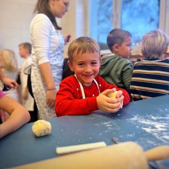 Eine Gruppe von Kindern und eine erwachsene Frau sind in einem Raum. Der Junge in Rot lächelt und hält Teig. Die Frau in Weiß steht hinter ihm. Die anderen Kinder beobachten. Es gibt einen Tisch mit einem Nudelholz und einer Schüssel mit Teig.