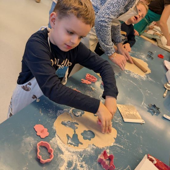 Ein Junge formt Teig mit Ausstechern, während ein anderes Kind zuschaut und ein Erwachsener hinter ihm hilft. Der Tisch ist mit Mehl und Ausstechern bedeckt.