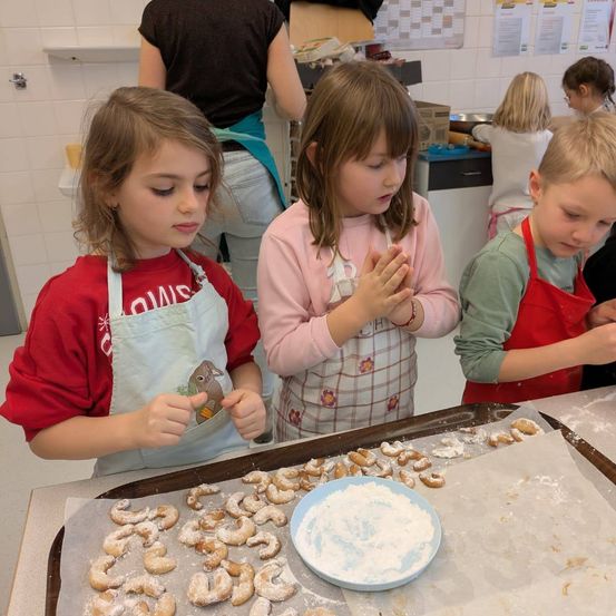Drei junge Kinder in Schürzen backen Kekse in einer Küche. Ein Mädchen in einem roten Shirt hält einen Keks. Ein Mädchen in einem rosa Shirt betet. Ein Junge in einem grünen Shirt schaut sich die Kekse an.