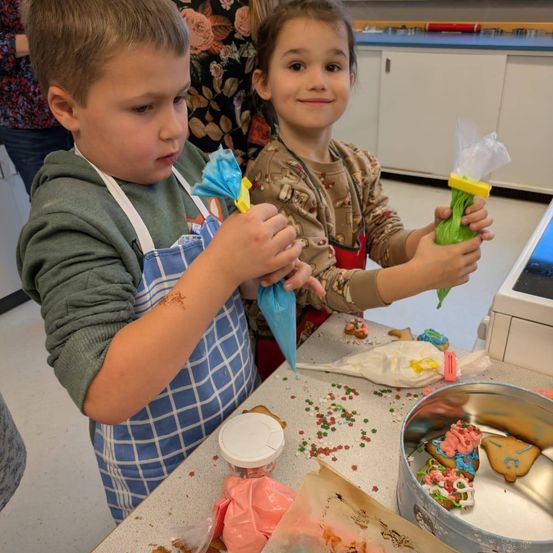 Zwei Kinder in einer Küche, ein Junge mit einer Schürze und ein Mädchen mit einer Teigtüte, dekorieren Kekse. In der Nähe gibt es einen Teller mit Keksen und eine Schüssel mit Zuckerguss. Hinter ihnen steht eine Person in der Nähe einer Theke mit Schränken.