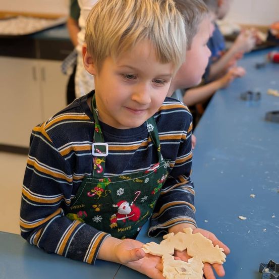 Ein junger Junge in einer Weihnachtsschürze lächelt, während er ausgeschnittene Lebkuchen in einer Klassenzimmerumgebung hält.