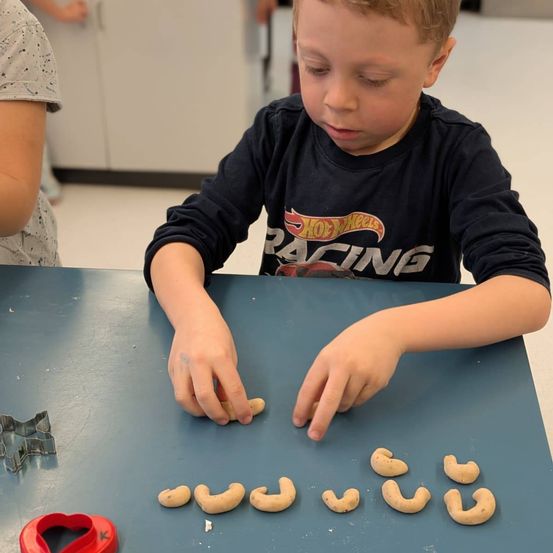 Ein junger Junge sitzt an einem Tisch und ordnet kleine, muschelförmige Objekte an. Er trägt ein schwarzes Shirt mit einem Design. Hinter ihm ist eine Person teilweise sichtbar. Werkzeuge wie Ausstechformen liegen auf dem Tisch.