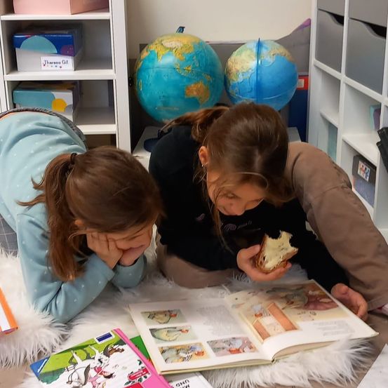 Zwei Mädchen sitzen auf dem Boden in einem Klassenzimmer und schauen in ein offenes Buch. Ein Mädchen hält ein Stück Essen. Dahinter sind zwei Globen und Regale mit Aufbewahrungsboxen zu sehen.