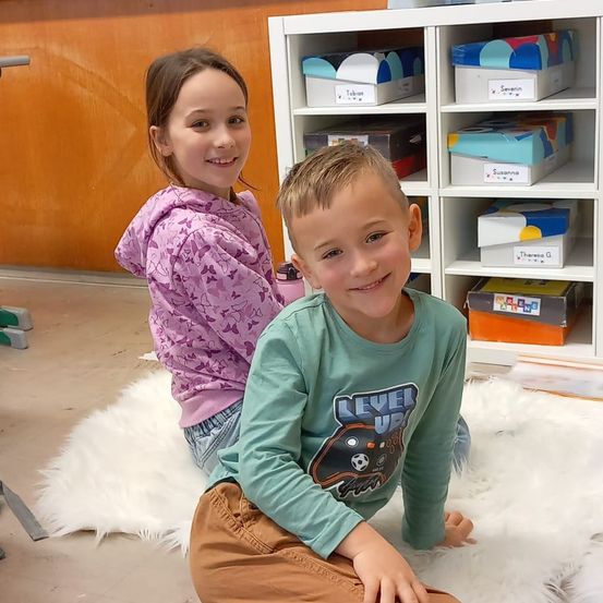 Ein Junge und ein Mädchen sitzen lächelnd auf einem weißen Pelzteppich. Sie befinden sich in einem Klassenzimmer mit Regalen, die mit Namen beschriftet sind.