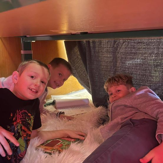 Drei junge Jungen liegen auf einem Bett, einer trägt ein schwarzes T-Shirt, ein anderer eine rosa Kapuzenjacke und der dritte ein weißes Hemd. Sie schauen nach oben.