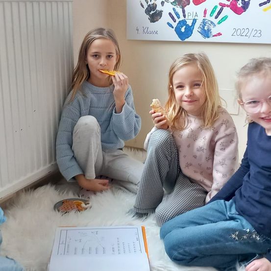 Drei Mädchen sitzen auf dem Boden und essen. Ein Mädchen isst eine Orangenscheibe, ein anderes hat einen Waffel und das dritte lächelt. Hinter ihnen ist ein weißer Teppich, ein Heizkörper und ein Brett mit Handabdrücken.