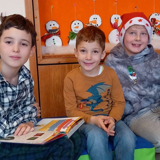 Drei Jungen sitzen auf einer Bank, einer liest ein Buch. Dahinter zeigt ein Hintergrund Schneemänner. Der Junge rechts trägt einen Weihnachtshut.