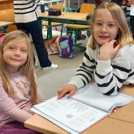 Zwei junge Mädchen sitzen an einem Schreibtisch in einem Klassenzimmer. Eine trägt eine Brille und die andere hat ein Einhorn auf ihrem Shirt. Sie lächeln und beschäftigen sich mit einem Buch vor ihnen.