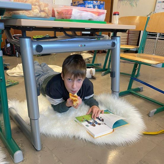 Ein junger Junge isst Pizza, während er auf einem weißen Pelzteppich unter einem Schreibtisch in einem Klassenzimmer liegt.