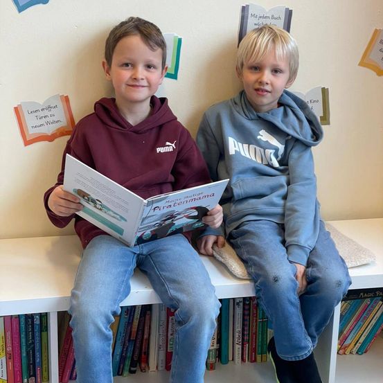Zwei junge Jungen sitzen auf einem Regal, einer liest ein Buch mit dem Titel 'Meine mutige Piratinnenmama' und der andere trägt ein Puma-Hoodie. Sie sind von Büchern auf Regalen umgeben.