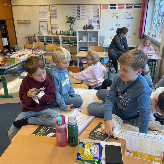In einem Klassenzimmer sitzen mehrere Kinder auf dem Boden und essen Snacks. Eine Frau steht hinter einem Schreibtisch. Bücher, Flaschen und Papiere liegen auf den Tischen. An der Wand stehen Pflanzen und Poster.