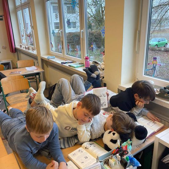 In einem Klassenzimmer liegen Kinder auf ihren Schreibtischen, lesen Bücher und ruhen sich aus. Ein Panda-Plüschtier und Scheren sind auf dem Schreibtisch. Draußen am Fenster ist ein grünes Auto geparkt.