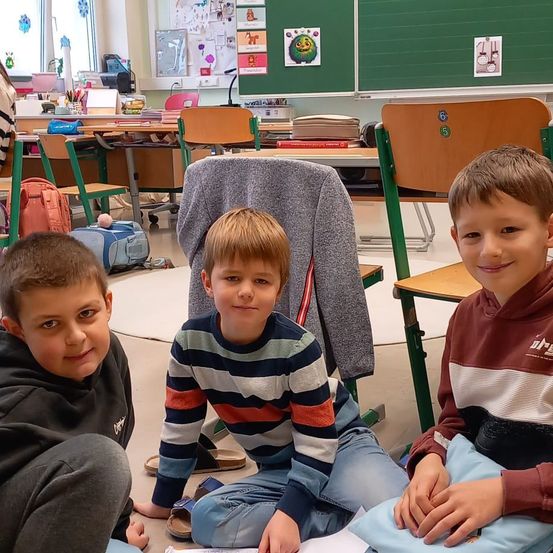 Drei Jungen sitzen lächelnd auf dem Boden in einem Klassenzimmer. Dahinter stehen Stühle, Tische und eine grüne Tafel. Ein grauer Pullover hängt an einem Stuhl. Ein Junge trägt ein gestreiftes Shirt.