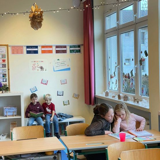 In einem Klassenzimmer sitzen zwei Jungen auf einer Bank und lesen Bücher. Vor ihnen sitzen zwei Mädchen an einem Schreibtisch mit einem rosa Becher und Büchern. Das Klassenzimmer hat eine weiße Wand mit bunten Papieren und einem Regal mit Büchern.