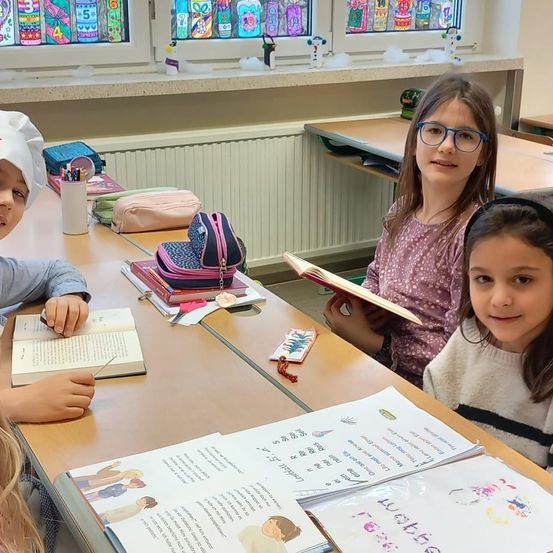 In einem Klassenzimmer sitzen drei Kinder an einem Tisch, jedes beschäftigt mit einem Buch. Ein Mädchen auf der rechten Seite liest, während ein Junge auf der linken Seite schreibt. Dahinter sind ein Heizkörper und farbenfrohe Fenster zu sehen.