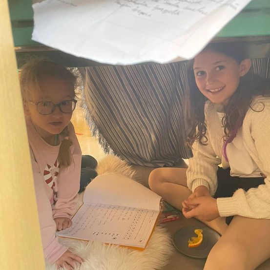 Zwei Mädchen sitzen in einer zeltartigen Struktur, eine mit Brille und einem Buch, die andere mit einem Teller Orange. Ein weißes Tuch ist über ihnen.