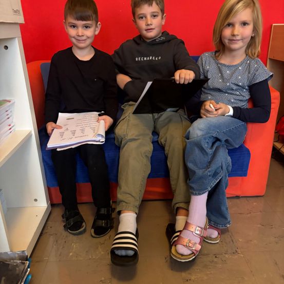 Drei Kinder sitzen lächelnd auf einem blauen Sofa. Der Junge links hält einen Stapel Papiere, der Junge in der Mitte hält ein Tablet, und das Mädchen rechts hält einen Stift. Sie sind in einem Raum mit einer roten Wand und weißen Regalen.