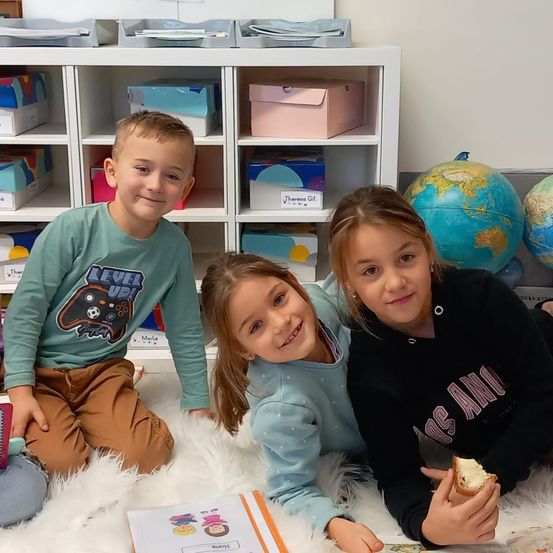 Drei Kinder sitzen auf einem weißen Teppich und lächeln für die Kamera. Ein Junge trägt ein grünes Shirt mit einem Videospiel-Design, während zwei Mädchen blaue und schwarze Sweatshirts tragen. Dahinter steht ein weißer Regal mit bunten Boxen und einer Erdkugel.