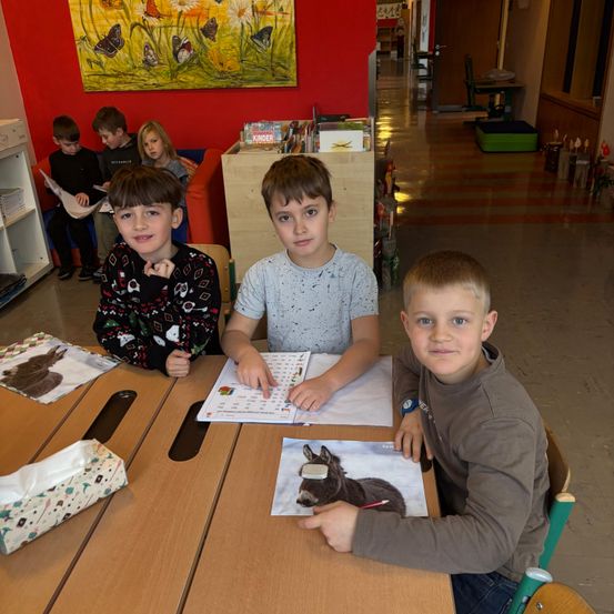 Vier Jungen sitzen an einem Tisch mit Büchern und Stiften in einem Klassenzimmer mit roter Wand. Ein Junge liest ein Buch, während andere ein offenes Buch und ein Eselbild betrachten.