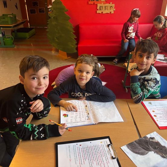 Drei Jungen sitzen mit Papieren und Bleistiften an einem Schreibtisch und lächeln für ein Foto. Dahinter ist ein Weihnachtsbaum und eine rote Couch mit Kindern zu sehen.
