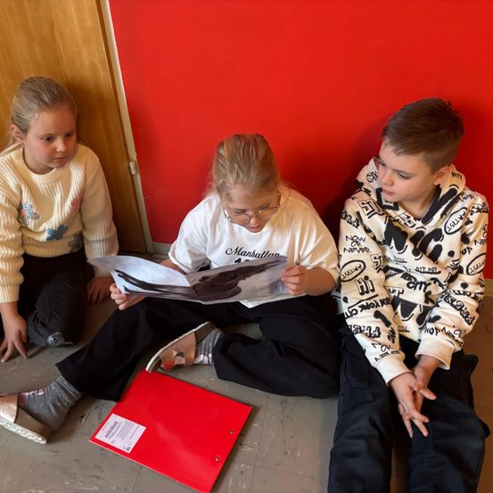 Drei Kinder sitzen auf dem Boden in der Nähe einer roten Wand. Eines der Mädchen liest aus einem Buch, während ein Junge und ein anderes Mädchen zusehen.