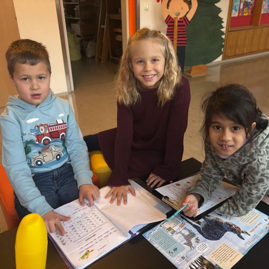 Drei Kinder sitzen an einem Tisch mit Büchern und einer gelben Flasche. Ein Mädchen lächelt, ein anderes schreibt und ein Junge schaut in ein Buch. Hinter ihnen hat eine Wand einen Weihnachtsbaum und eine Kinderzeichnung.
