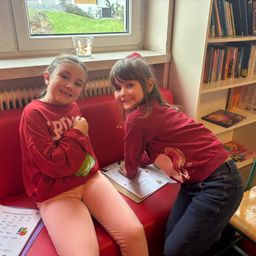 Zwei junge Mädchen sitzen auf einem roten Sofa und lächeln. Eines hält eine Klemmbrett mit einem Formular und das andere einen Stift. Dahinter steht ein Bücherregal voller Bücher.