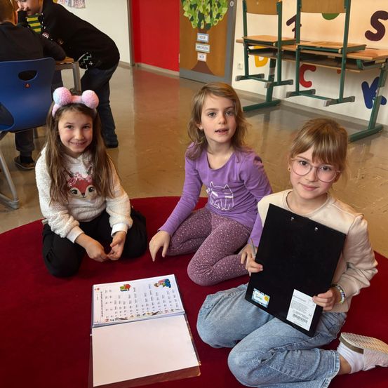 Drei junge Mädchen sitzen auf einem roten Teppich und lächeln für ein Foto. Sie halten ein Buch und eine Mappe. Eine von ihnen hat eine Kopfbedeckung mit Ohren. Hinter ihnen sind Stühle und ein Tisch. Ein Junge steht im Hintergrund.